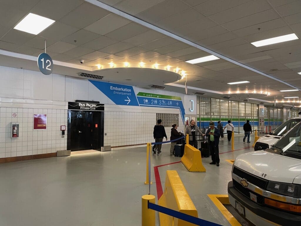 The image shows an indoor area resembling a transportation terminal or parking garage. There are several people with luggage standing near a row of parked white vans. Overhead signs indicate directions for embarkation, with numbers and letters such as "12" and "J" visible. A door labeled "Pan Pacific Fitness Club" is on the left, and the area is well-lit with ceiling lights. The floor has yellow barriers and red lines, suggesting designated pathways or parking spaces.