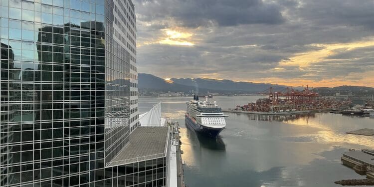 The image shows a large cruise ship approaching a dock in a harbor. To the left, there is a tall glass building reflecting the surrounding area. In the background, there are cranes and shipping containers on a dock, with mountains and a cloudy sky at sunset. The water reflects the colors of the sky, creating a serene atmosphere.