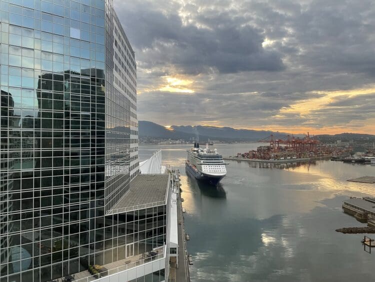 The image shows a large cruise ship approaching a dock in a harbor. To the left, there is a tall glass building reflecting the surrounding area. In the background, there are cranes and shipping containers on a dock, with mountains and a cloudy sky at sunset. The water reflects the colors of the sky, creating a serene atmosphere.