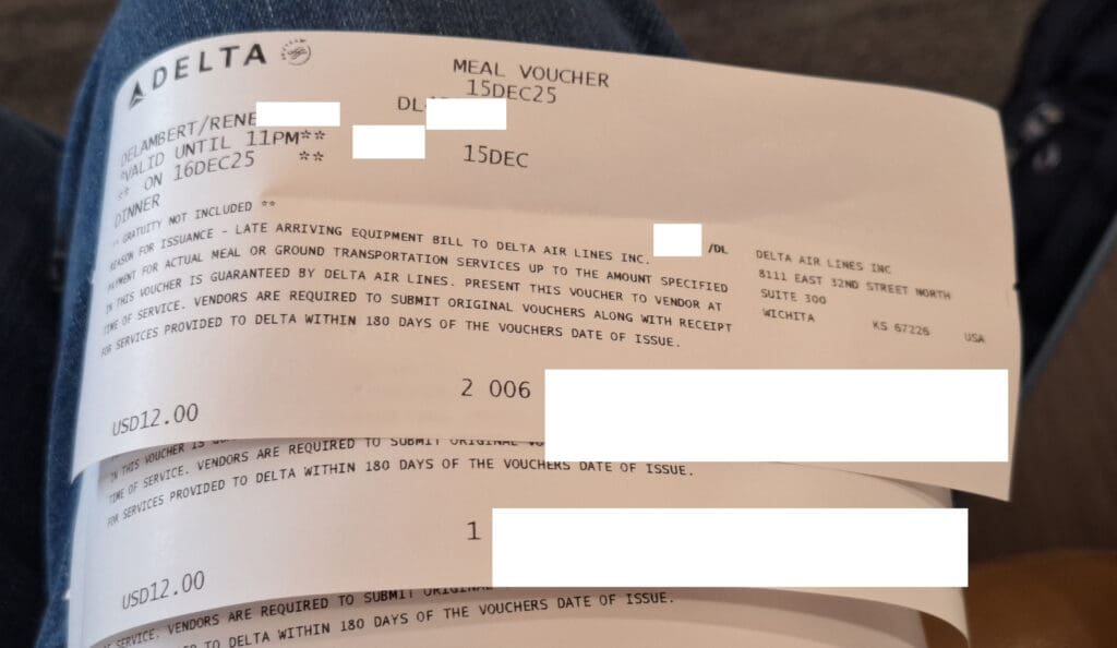 The image shows a Delta Air Lines meal voucher. It includes details such as the date "15DEC25," a validity time until 11 PM on "16DEC25," and a note that gratuity is not included. The voucher is for a dinner valued at USD 12.00. It mentions that the voucher is issued due to late-arriving equipment and is to be billed to Delta Air Lines. The voucher includes instructions for vendors to submit original vouchers with receipts within 180 days of the issue date. Some personal information is redacted.