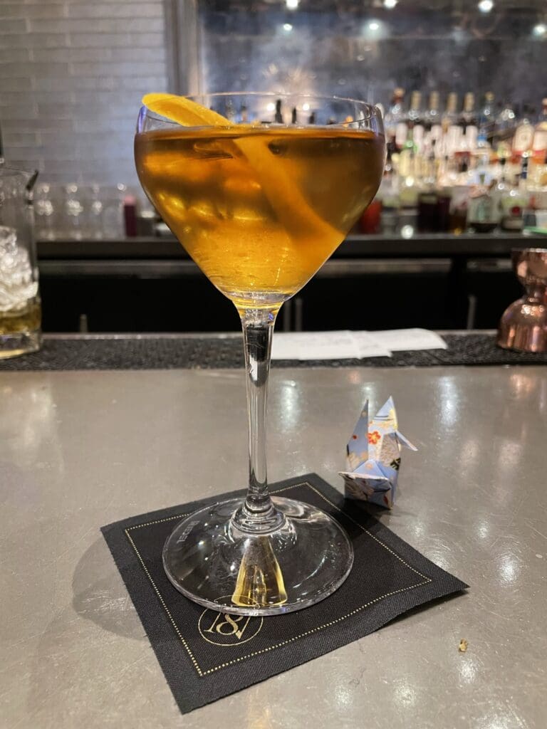 A cocktail glass filled with an amber-colored drink, garnished with an orange twist, is placed on a black napkin on a bar counter. In the background, there are blurred bottles on shelves. Next to the glass, there is a small origami figure.