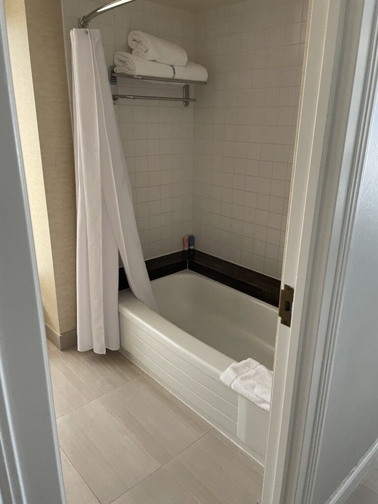 The image shows a bathroom with a white bathtub and a shower curtain partially pulled to the side. Above the tub, there is a metal shelf holding neatly folded white towels. The walls are tiled, and there are small bottles of toiletries on the edge of the tub. A white towel is draped over the side of the bathtub. The floor is covered with light-colored tiles.