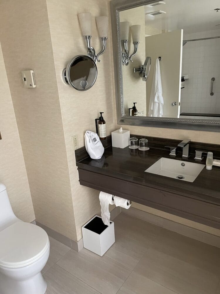 The image shows a bathroom with a modern design. There is a toilet on the left and a countertop with a sink on the right. Above the sink is a large mirror with two wall-mounted light fixtures. A magnifying mirror is attached to the wall. On the countertop, there are toiletries, a tissue box, and two glasses. Below the counter, a roll of toilet paper is hanging, and a small waste bin is on the floor. A towel is hanging on the door, which is slightly open, revealing a shower area with a grab bar.