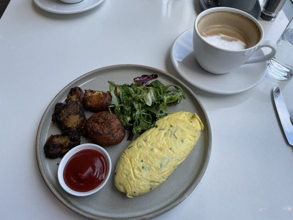 A plate of food is on a table, featuring an omelette, a small salad, and crispy roasted potatoes with a small dish of ketchup. Next to the plate is a cup of coffee with foam on top, placed on a saucer. A glass of water and a knife are also visible on the table.