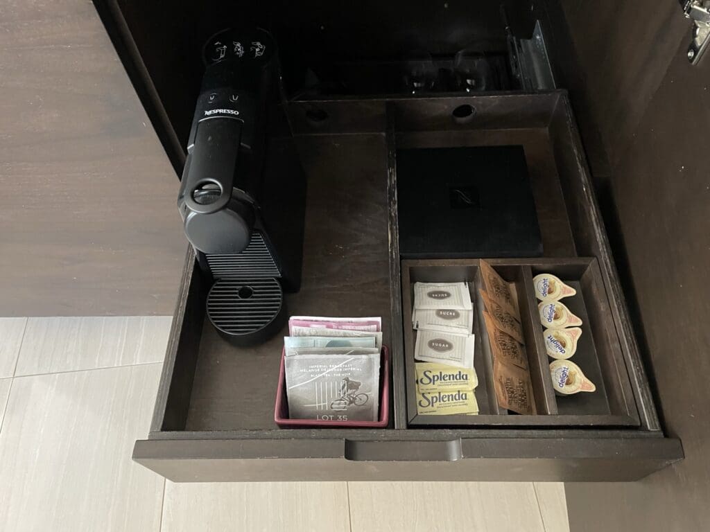 The image shows an open drawer containing a Nespresso coffee machine on the left. Next to it, there are compartments holding various items: tea bags, sugar packets, Splenda sweetener packets, coffee pods, and creamers. The drawer is part of a dark wooden cabinet, and the floor is light-colored tile.