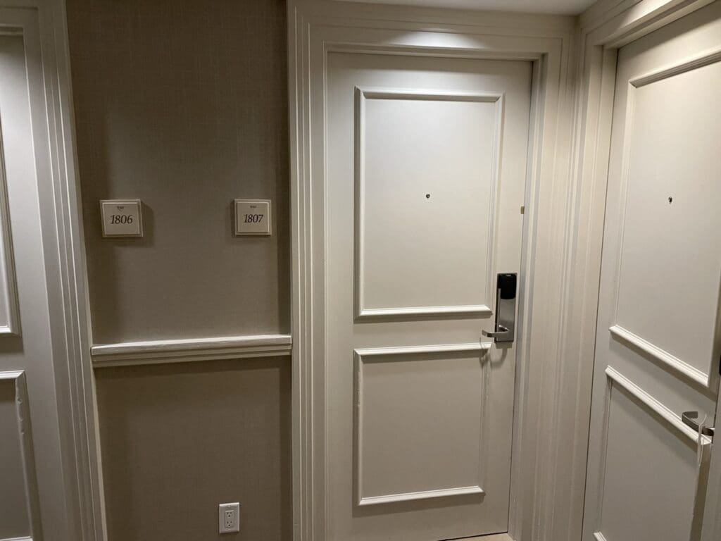 The image shows a hallway in a hotel or apartment building with three doors. The door in the center has a room number sign next to it labeled "1807," and the door to the left has a sign labeled "1806." Each door has a peephole and a modern electronic lock. The walls are light-colored, and there is a small electrical outlet visible near the floor.