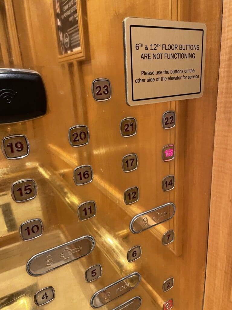 The image shows an elevator control panel with buttons for different floors. A sign on the panel indicates that the 6th and 12th floor buttons are not functioning and advises using buttons on the other side of the elevator for service. The panel has a metallic finish, and the buttons are arranged in a grid.