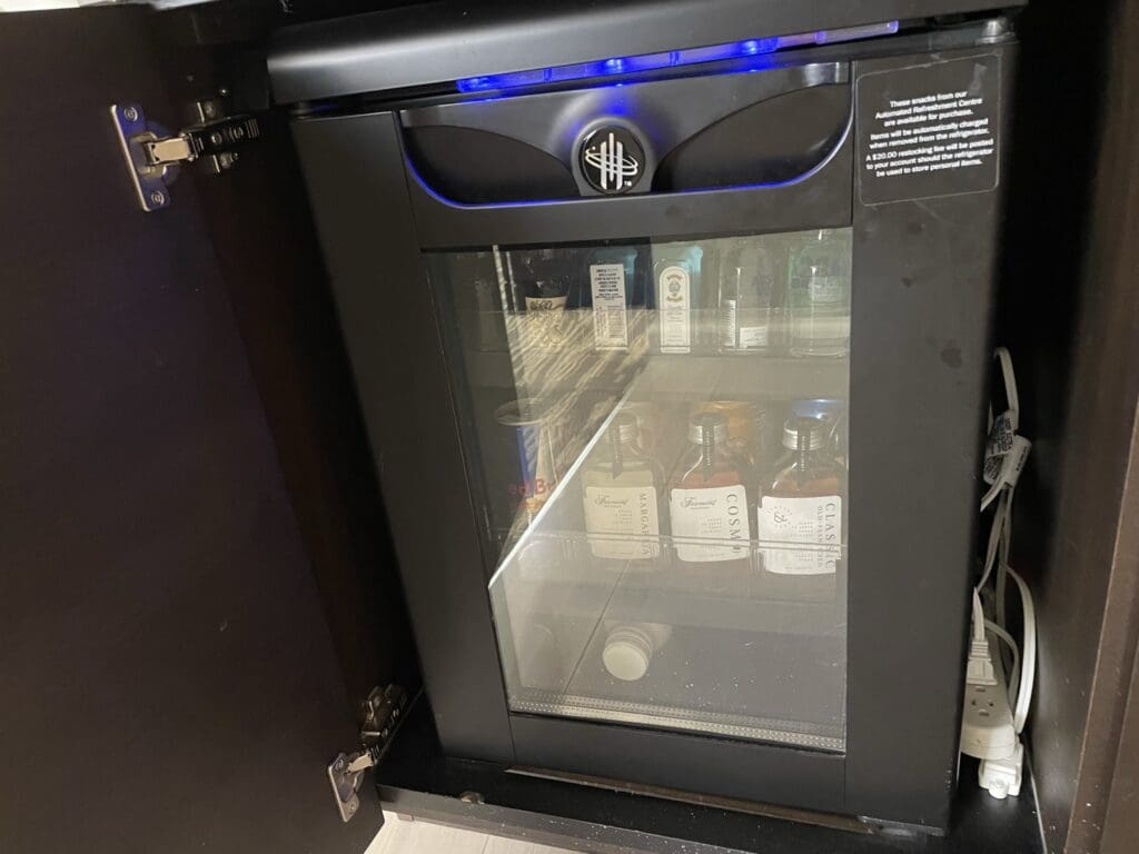 The image shows a small hotel minibar fridge with a glass door, partially open in a cabinet. Inside, there are various beverages, including small bottles of alcohol and cans. A sign on the fridge door provides information about charges for items removed. The fridge is plugged into a power strip visible to the right.