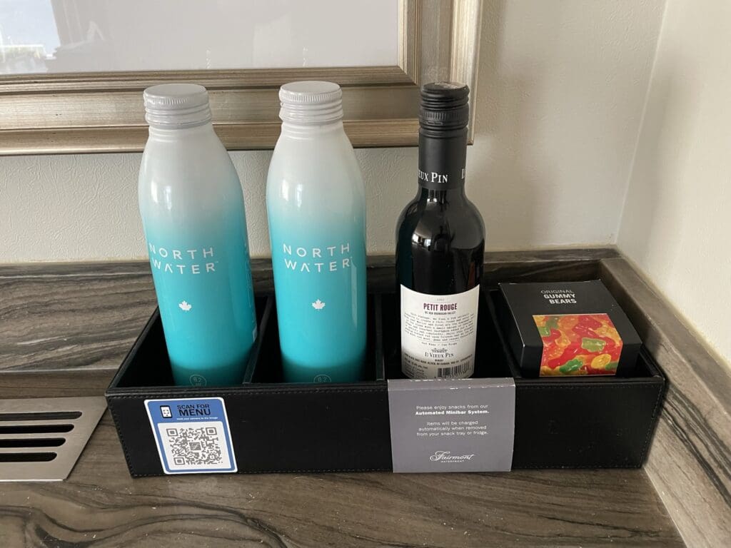 The image shows a minibar setup on a countertop. It includes two bottles of "North Water" with a gradient blue and white design, a bottle of red wine labeled "Petit Rouge," and a box of "Original Gummy Bears." These items are placed in a black tray. There is also a QR code on the tray labeled "Scan for Menu," and a note about the automated minibar system.