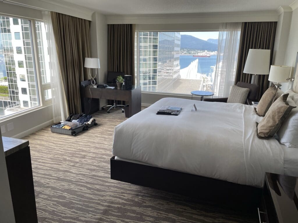 A hotel room with a large bed covered in white linens and several pillows. There is a desk with a chair, a lamp, and some electronic devices near the window. An open suitcase with clothes is on the floor. The room has large windows with a view of a cityscape, including tall buildings, water, and mountains in the background. The carpet has a patterned design, and there are curtains on the windows.