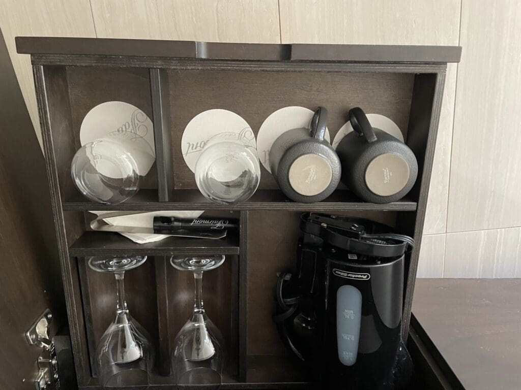 The image shows a wooden cabinet with various drinkware and a small appliance. On the top shelf, there are two upside-down glass cups and two upside-down black mugs, each placed on a coaster. Below, there are two upside-down wine glasses. A napkin and a pen are also visible. On the right side, there is a black electric kettle.