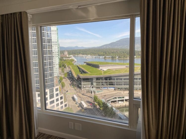 View from a window showing a cityscape with tall buildings, a green-roofed structure, and a waterfront. In the background, there are mountains under a clear blue sky. The window is framed by brown curtains.
