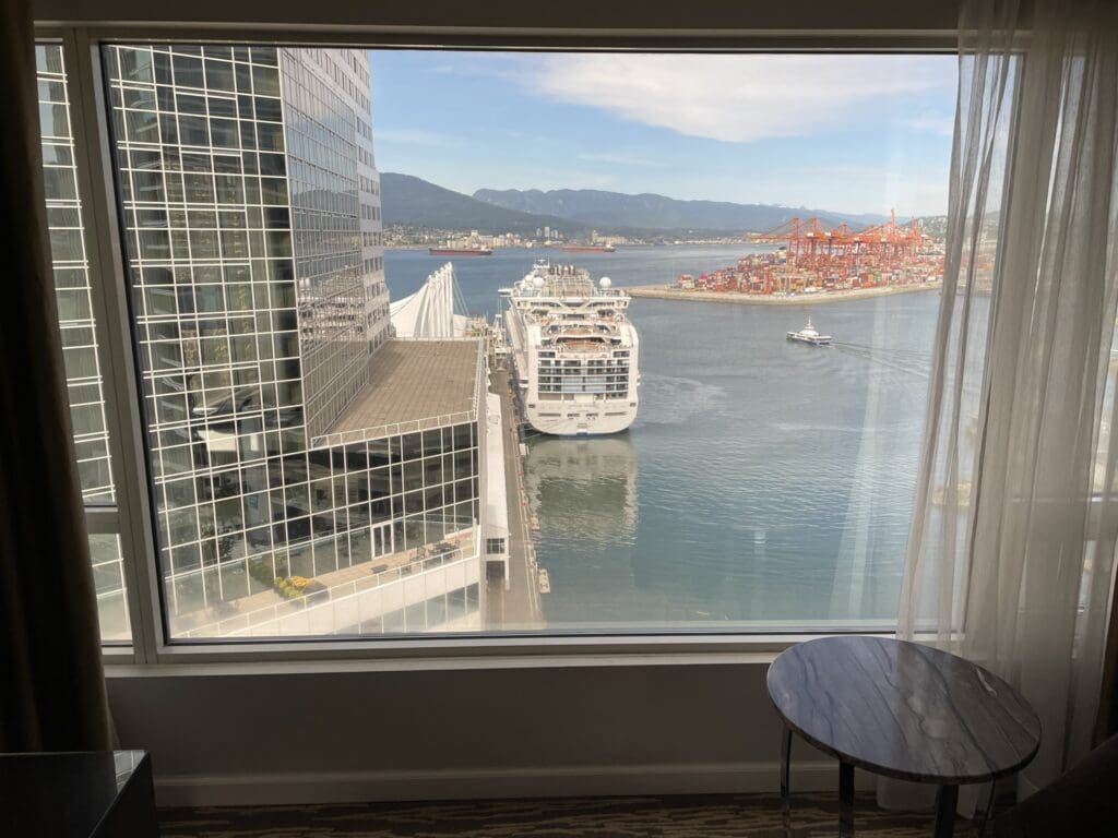 The image shows a view from a window overlooking a harbor. A large cruise ship is docked next to a modern glass building. In the background, there are mountains and a port with cranes and shipping containers. A small round table is visible in the foreground by the window.