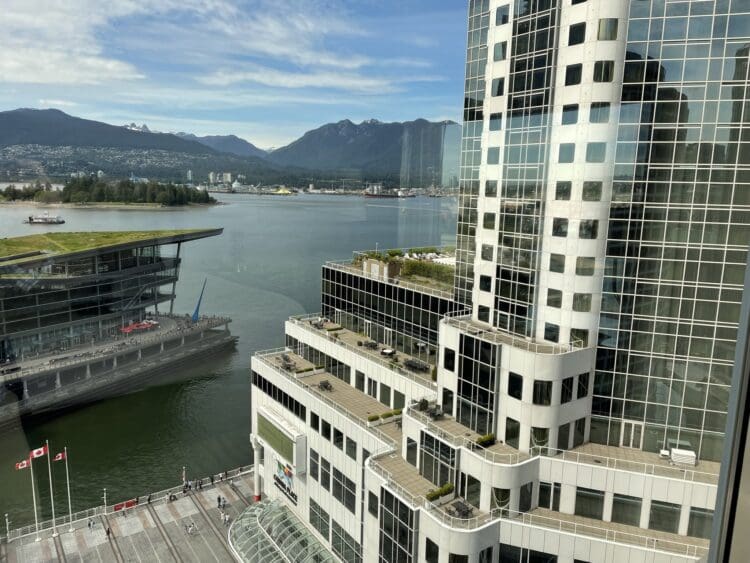 A view from a high vantage point showing a waterfront scene. In the foreground, there is a modern building with a glass facade and multiple terraces. To the left, a building with a green roof is visible, adjacent to the water. In the background, there are mountains under a partly cloudy sky. The waterway has a few boats, and there are Canadian flags on poles near the bottom of the image.