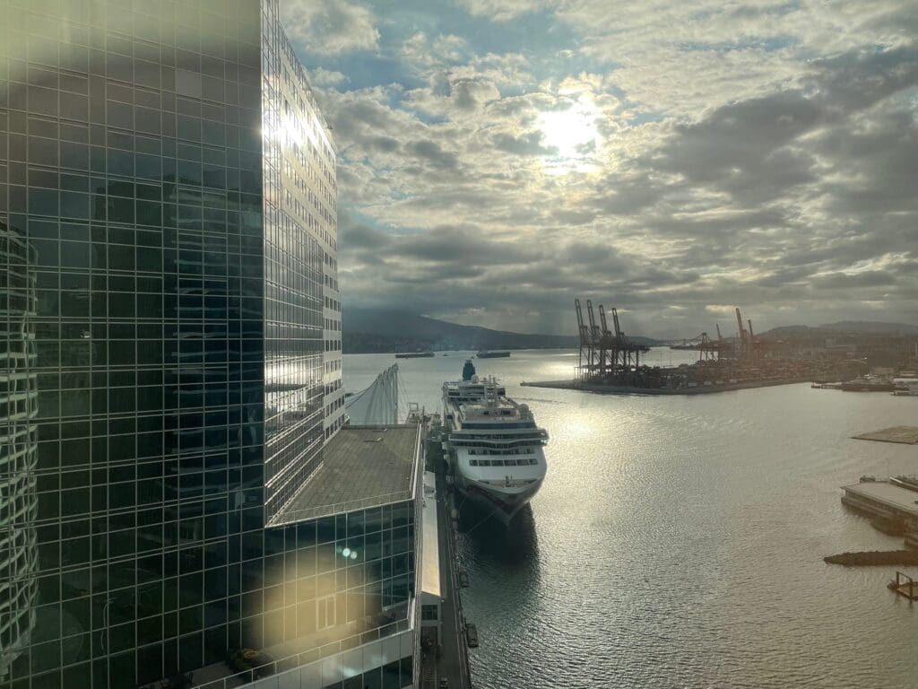 A large cruise ship is docked at a port next to a modern glass building. The sun is partially obscured by clouds, casting reflections on the water. In the background, there are several cranes and industrial structures along the waterfront. The scene conveys a busy harbor area with a mix of urban and industrial elements.