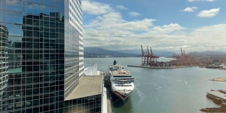 A large cruise ship is docked at a port next to a modern glass building. The building reflects the surrounding area, including the water and sky. In the background, there are several red cranes and containers at a shipping terminal. The sky is partly cloudy, and there are mountains visible in the distance.