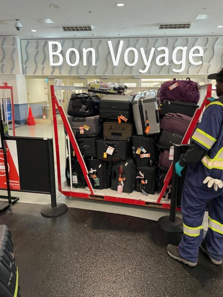 A large stack of various suitcases and bags is piled on a cart in an airport or travel setting. A person in a blue uniform with reflective stripes stands nearby. Above the luggage, a sign reads "Bon Voyage."