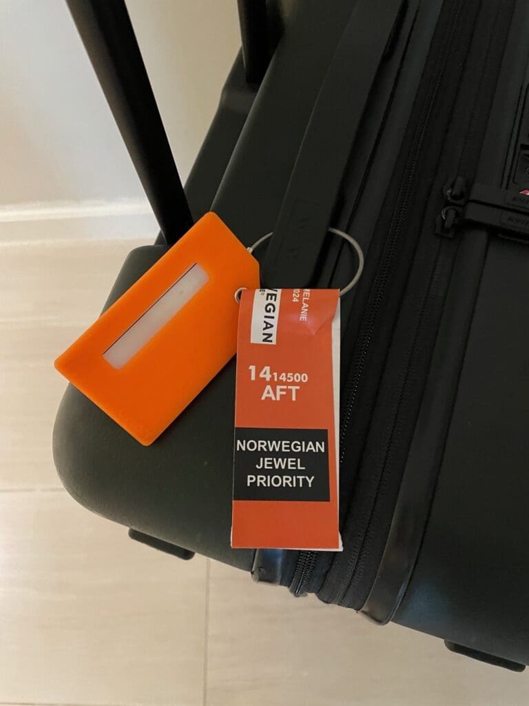A black suitcase with an orange luggage tag and a red and white priority tag attached. The tag reads "NORWEGIAN JEWEL PRIORITY" and includes the text "14 AFT." The suitcase is positioned on a light-colored floor.