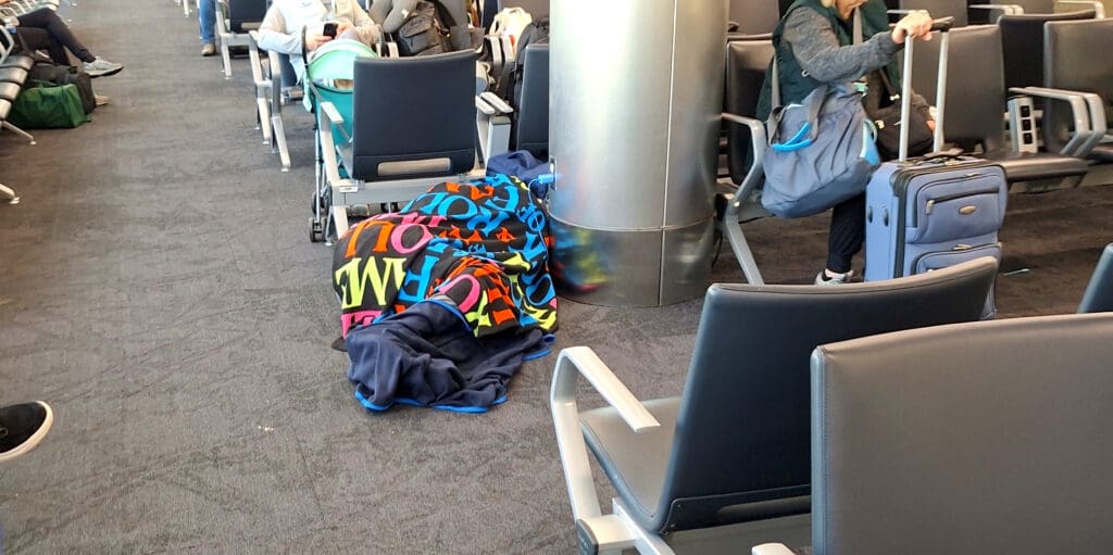 A person is lying on the floor of an airport terminal, covered with a colorful blanket featuring large letters. They are next to a metal column. Nearby, people are seated in chairs, and a woman is pulling a suitcase. The area is filled with rows of chairs and travelers.
