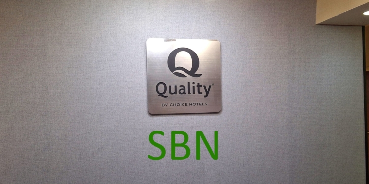 The image shows a wall with a metallic sign displaying the logo and text "Quality by Choice Hotels." Below the sign, the letters "SBN" are written in green. The wall has a textured surface, and there is a ceiling light visible in the top right corner.