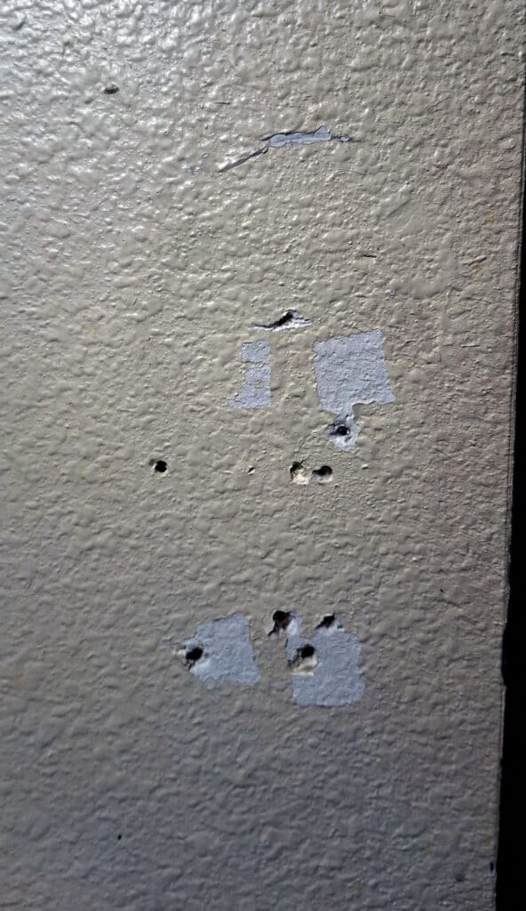 A close-up of a textured wall with several small holes and patches of peeling paint. The wall is painted in a light color, and the exposed areas reveal a darker layer underneath.