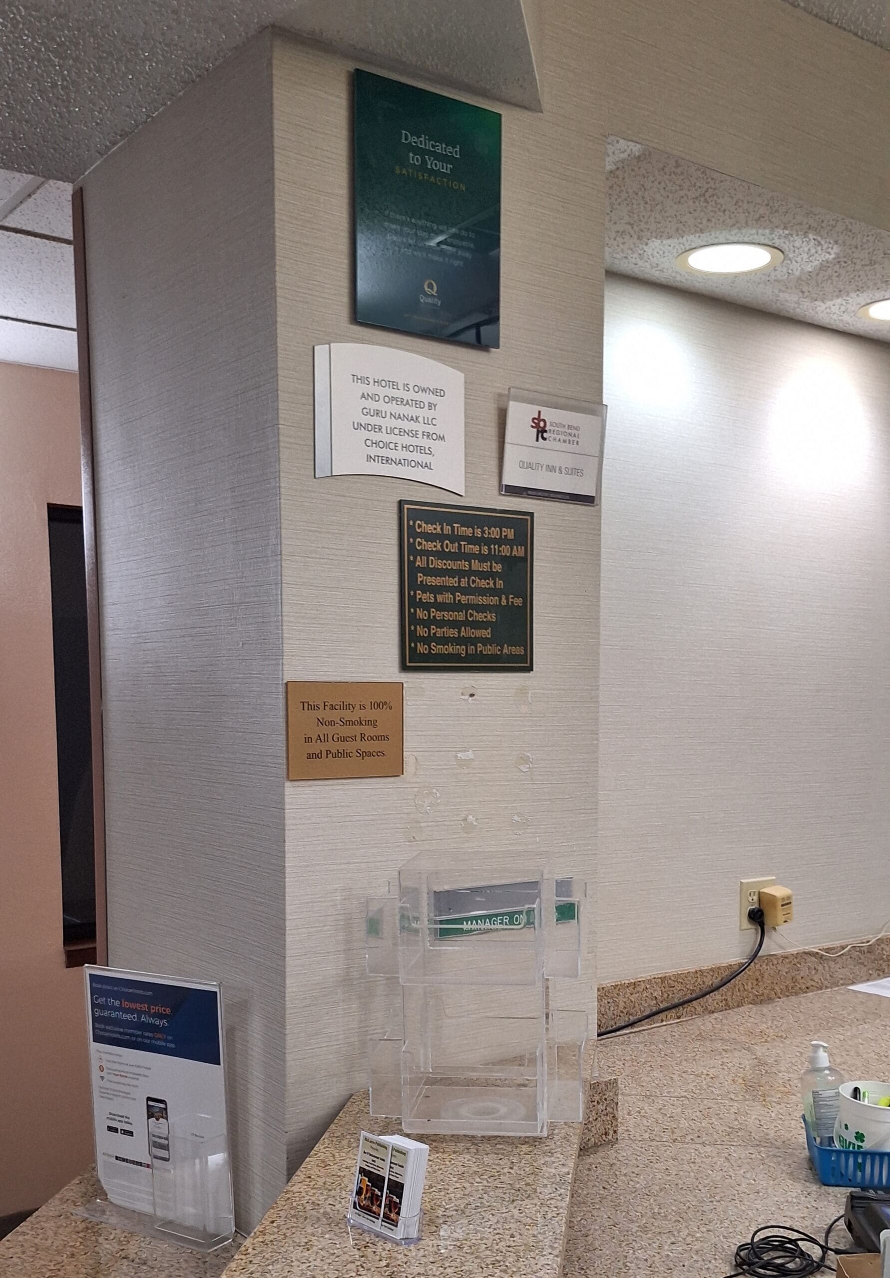 The image shows a hotel reception area with several signs on the wall. The signs include information about hotel ownership, check-in and check-out times, and policies such as no smoking and pet fees. There is a clear plastic brochure holder on the counter, along with a small basket containing hand sanitizer and other items. The wall and counter are light-colored, and there is a power outlet with a plug on the counter.