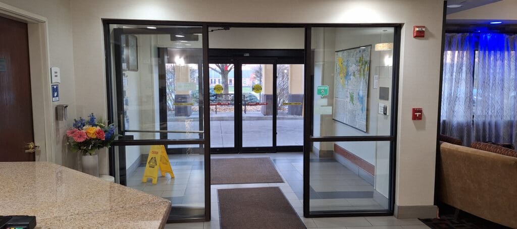 The image shows the interior of a building entrance with glass doors leading outside. There is a yellow caution sign on the floor, a bouquet of colorful flowers in a vase on the left, and a map on the wall to the right. The area is well-lit, with a granite countertop in the foreground and a fire alarm on the wall.