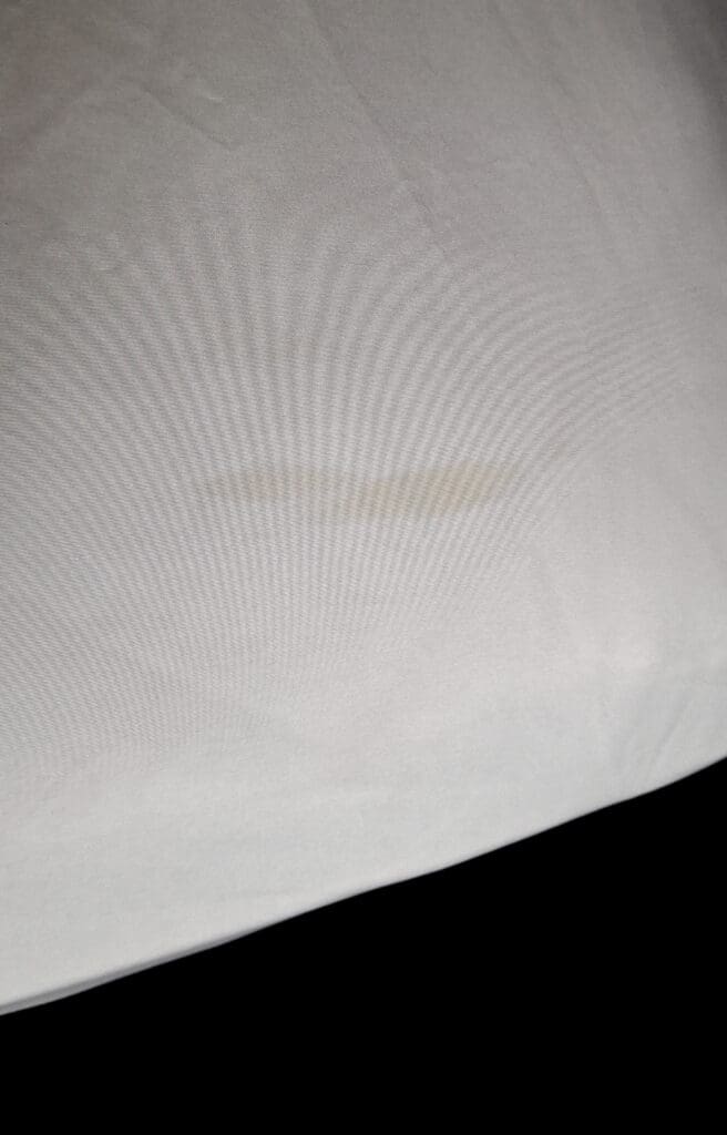 The image shows a close-up of a light-colored fabric with a faint, elongated stain in the center. The background is plain and the fabric appears to be smooth.