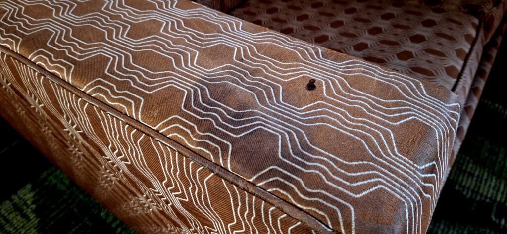 The image shows a close-up of a brown upholstered chair with a geometric pattern. The fabric features a series of white, wavy lines forming hexagonal shapes. There is a small dark spot on the fabric.