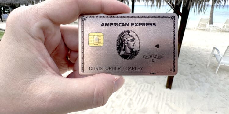 A hand is holding an American Express credit card with a beach setting in the background. The card features a chip and contactless symbol, and there are thatched umbrellas and lounge chairs on the sandy beach.