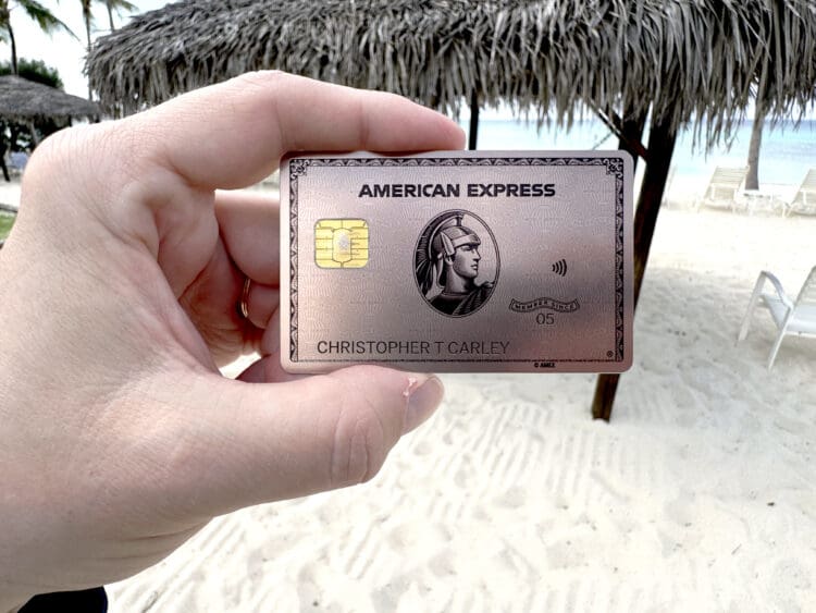 A hand is holding an American Express credit card with a beach setting in the background. The card features a chip and contactless symbol, and there are thatched umbrellas and lounge chairs on the sandy beach.