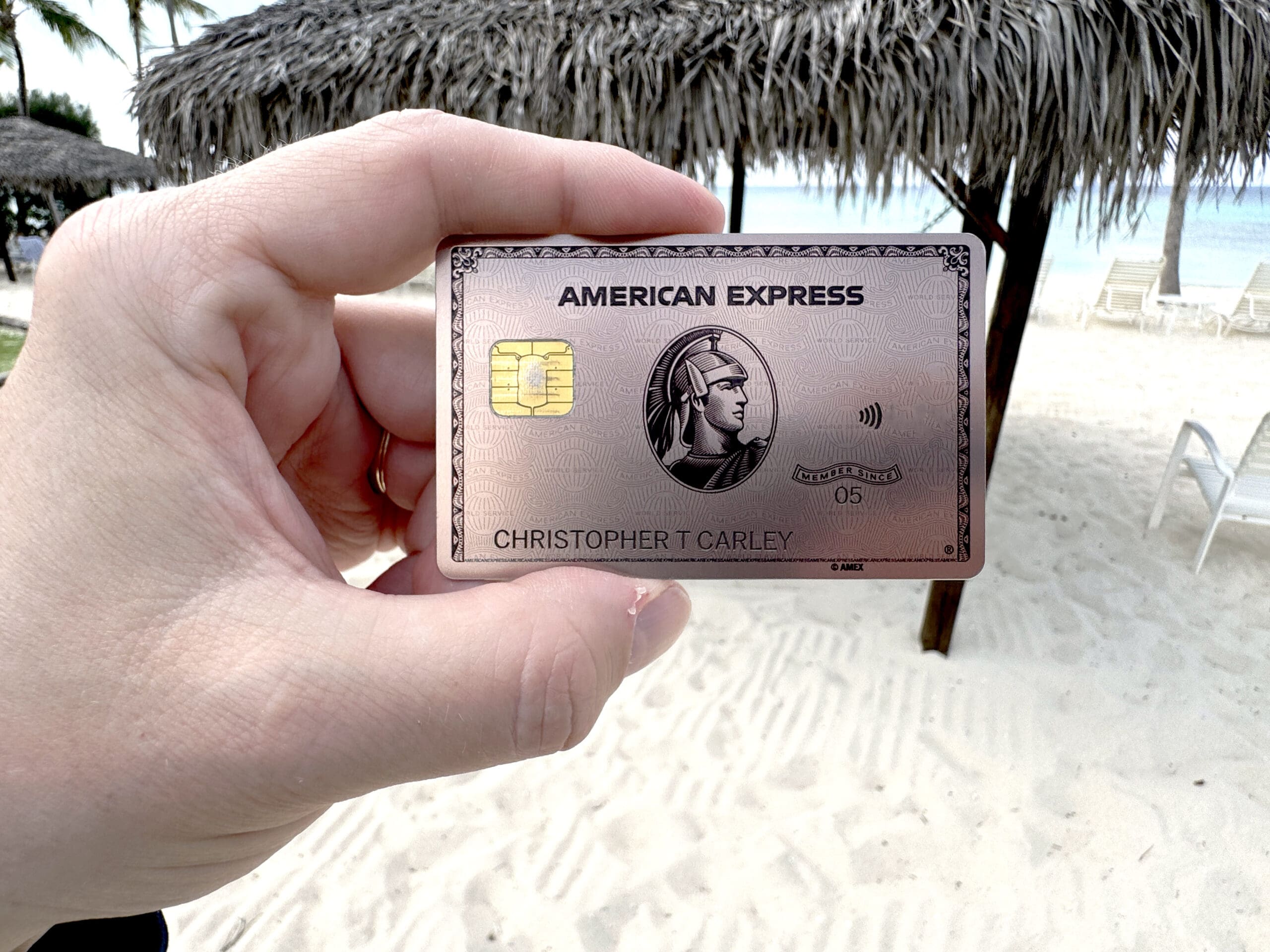 A hand is holding an American Express credit card with a beach setting in the background. The card features a chip and contactless symbol, and there are thatched umbrellas and lounge chairs on the sandy beach.