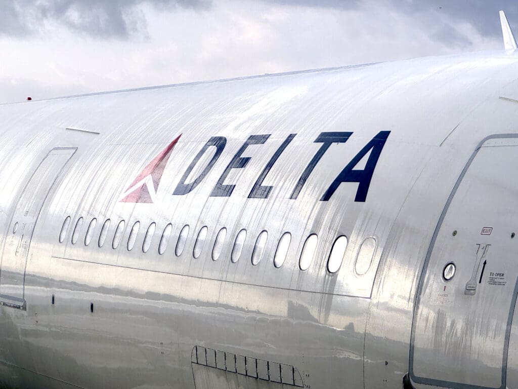 The image shows a close-up of the side of a Delta Air Lines airplane. The aircraft's metallic body is visible, with the Delta logo and name prominently displayed. There are several windows along the side, and the sky is overcast in the background. A Delta Air Lines A330-300 (©Eye of the Flyer)