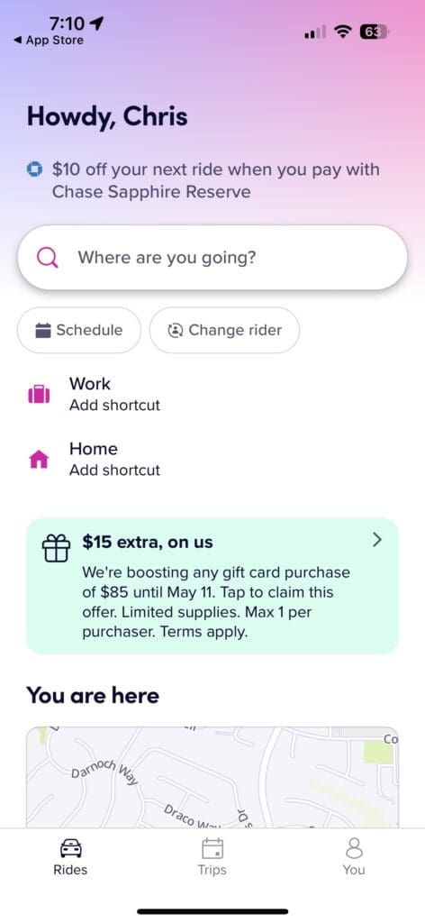 The image is a screenshot of a ride-sharing app interface. It greets the user with "Howdy, Chris" and offers a $10 discount on the next ride with Chase Sapphire Reserve®. There is a search bar asking "Where are you going?" with options to schedule a ride or change the rider. Shortcuts for "Work" and "Home" can be added. A promotional offer of "$15 extra, on us" for gift card purchases is displayed. A small map section shows the user's current location. At the bottom, there are navigation icons for "Rides," "Trips," and "You."