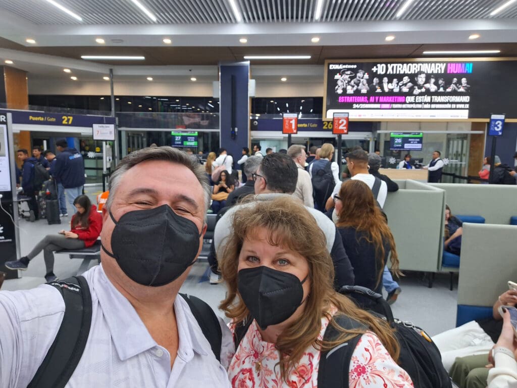A man and a woman wearing black face masks are taking a selfie in an airport terminal. They are surrounded by other travelers, some seated and some standing in line. Behind them, there are signs for gates 26 and 27, and a large advertisement on the wall. The terminal is well-lit with modern seating and electronic displays.