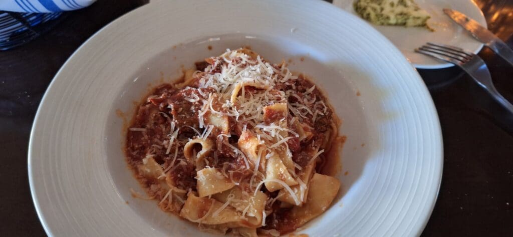 A plate of pasta with a rich tomato and meat sauce, topped with grated cheese. The pasta appears to be pappardelle. In the background, there is a small plate with a piece of bread and a fork and knife at Parisi restaurant in South Bend, Indiana. Parisi is a Resy restaurant. 
