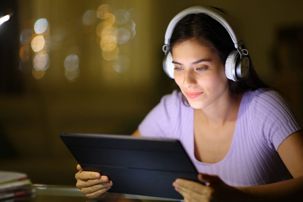 A woman wearing headphones is looking at a tablet. She is sitting indoors with a soft focus background featuring warm, blurred lights. She appears to be engaged and focused on the screen.