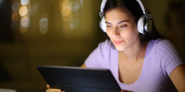 A woman wearing headphones is looking at a tablet. She is sitting indoors with a soft focus background featuring warm, blurred lights. She appears to be engaged and focused on the screen.