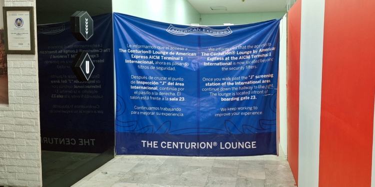 The image shows a hallway with a large blue banner displaying information about the location of The Centurion® Lounge by American Express at AICM Terminal 1 International. The text is in both Spanish and English, indicating that the lounge is now located beyond the security filters, near boarding gate 23. The hallway has a red and white wall on the right and a framed certificate on the left. There are also two illuminated signs with symbols above the banner.