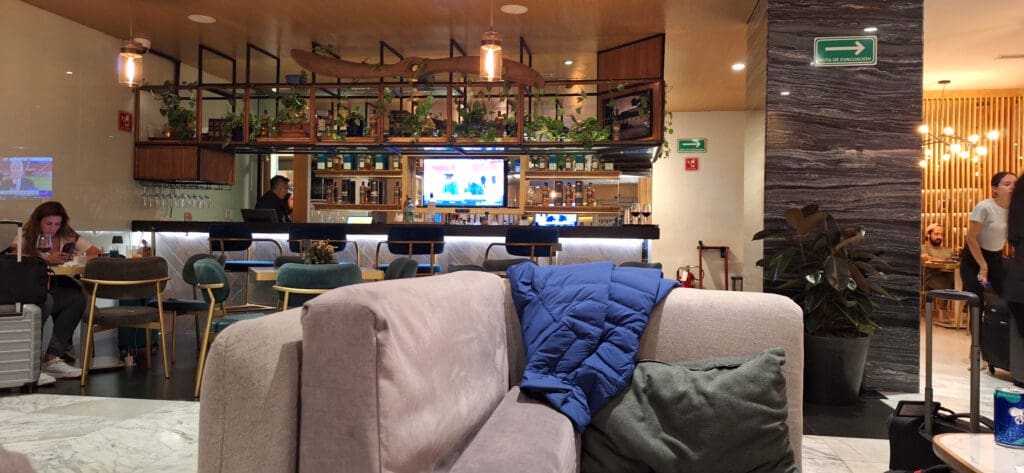 The image shows a cozy lounge area with a bar in the background. There are several chairs and tables, with a few people sitting and engaging in various activities. A woman is seated on the left, looking at her phone with a drink on the table. A blue jacket is draped over a beige sofa in the foreground. The bar is well-stocked with bottles and has a TV screen displaying a program. Green plants are placed on a shelf above the bar. There are signs on the walls, including an emergency exit sign. The lighting is warm and inviting.