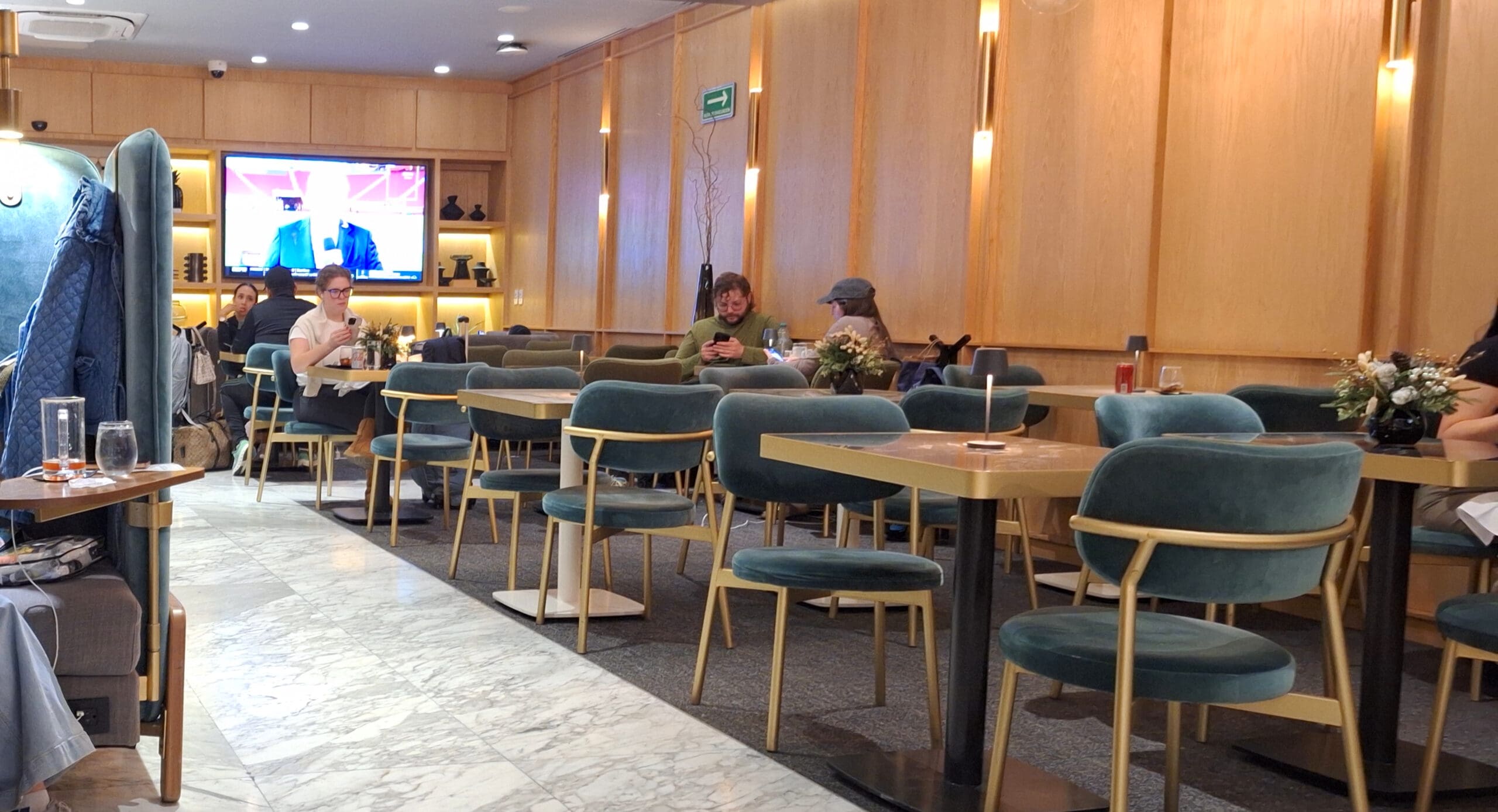 A modern cafe interior with several tables and teal chairs. People are seated at some tables, engaged in conversation or using their phones. A large TV screen on the wall displays a person speaking. The decor includes wooden paneling, soft lighting, and floral arrangements on the tables. An exit sign is visible on the wall.
