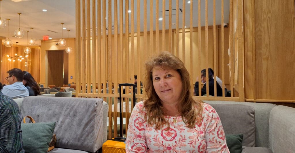 A woman with long hair is sitting on a gray couch in a modern, warmly lit room with wooden decor. She is wearing a white and pink patterned top and smiling at the camera. In the background, there are other people sitting and talking, with hanging spherical lights and wooden partitions adding to the ambiance. A suitcase is visible next to the couch.