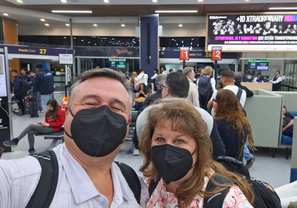 A man and a woman wearing black face masks are taking a selfie in an airport terminal. They are surrounded by other travelers, some seated and some standing in line. Behind them, there are signs for gates 26 and 27, and a digital billboard displaying an advertisement. The atmosphere is busy, with people waiting and moving around.