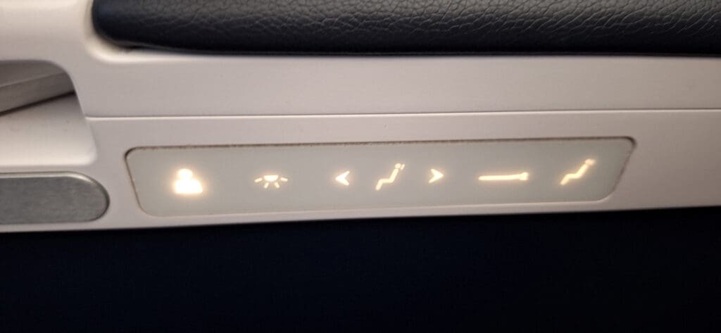 The image shows a close-up of an airplane seat control panel. It features illuminated icons for various seat adjustments, including a person icon, a light, and arrows indicating seat recline and leg rest positions. The panel is set into the armrest of the seat.