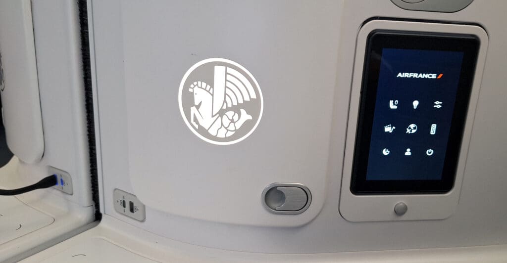 The image shows a section of an airplane seat with a control panel and a logo. The control panel displays the Air France logo and various icons for seat and flight controls. Below the panel, there are USB ports with a cable plugged into one of them. On the left, there is a circular logo featuring a stylized horse with wings and a globe.