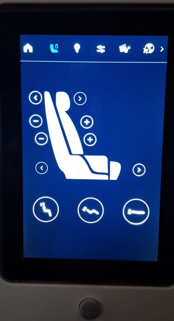The image shows a digital control panel for adjusting a seat, likely in a vehicle or airplane. The screen displays a diagram of a seat with various adjustment options, including buttons for moving the seat forward or backward, and increasing or decreasing lumbar support. There are icons at the top for different functions, such as a home button, phone, light, and others. At the bottom, there are icons representing different seating positions.