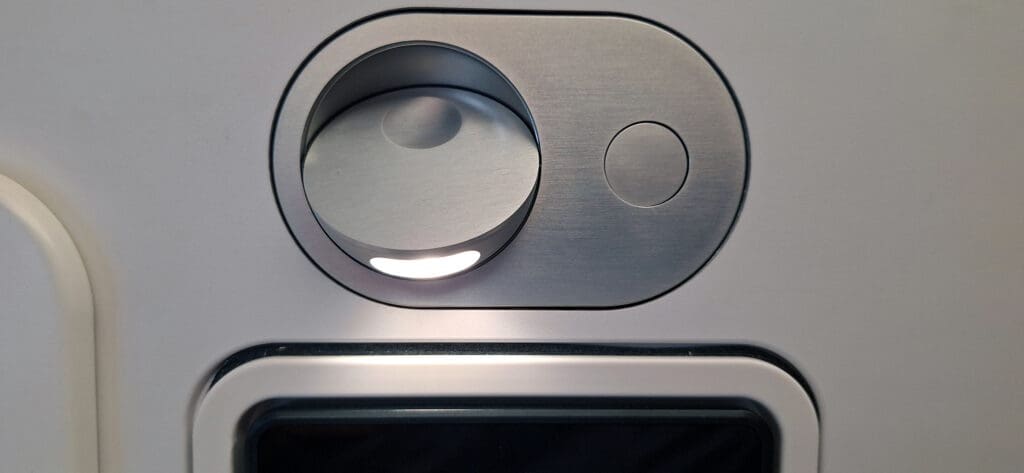 The image shows a close-up of an airplane cabin light and air vent control panel. The panel is metallic with a circular air vent on the left and a smaller circular button on the right. Below the panel, part of a screen or display is visible.