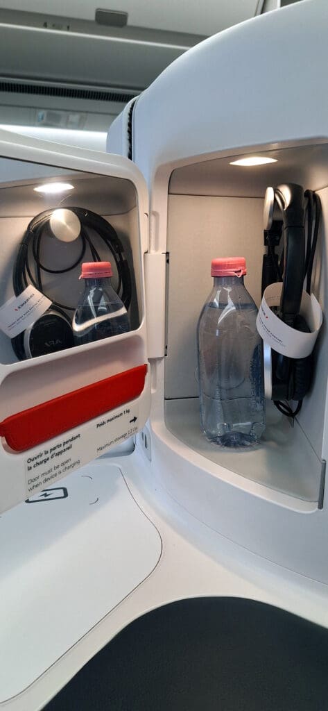 The image shows an open storage compartment in an airplane, containing two water bottles with pink caps and two sets of headphones. The compartment has a small light illuminating the contents. There is a red strap and a label with instructions in both French and English about opening the door while charging a device.