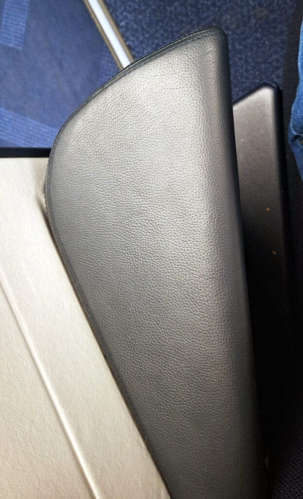 The image shows a close-up of an airplane seat armrest. The armrest is covered in a textured, dark gray material. The background includes parts of the airplane's interior, such as the floor and another seat.
