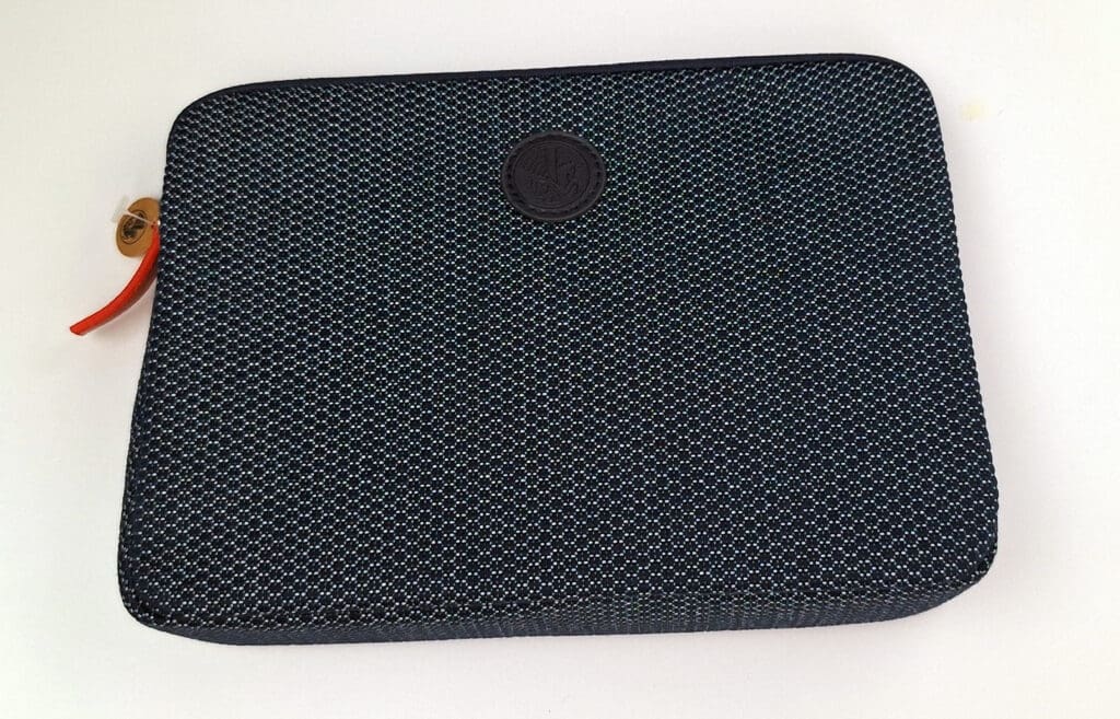 This image shows a rectangular, textured fabric case with a dark color and a subtle pattern. It has a circular emblem in the center and a red zipper pull on the left side. The case is placed on a white surface.